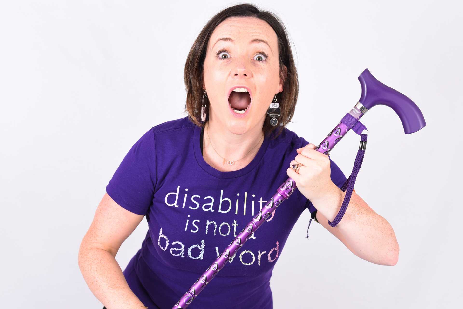 Lisette in a purple t-shirt which reads ‘disability is not a bad word’, holding her purple walking stick with her eyes and mouth wide open