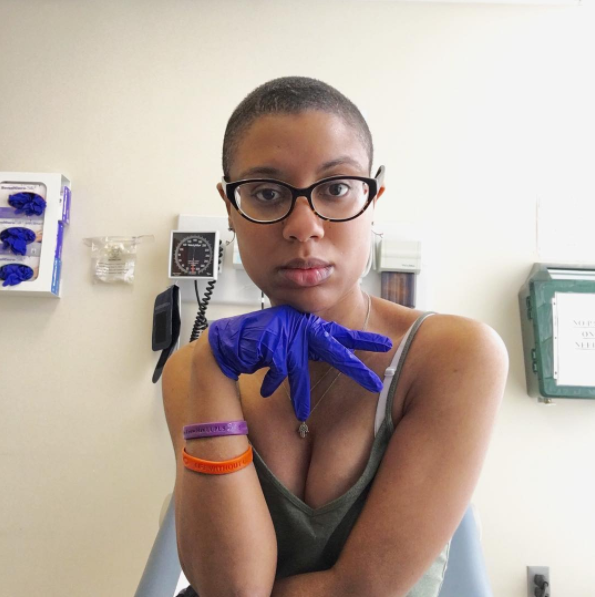 A woman with cropped hair and glasses sits in hospital room wearing blue surgical gloves. She poses for the camera