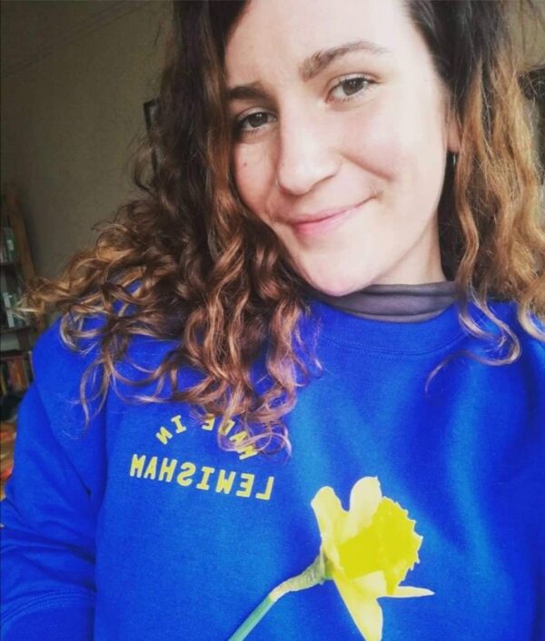 Lauren smiles at the camera holding a yellow daffodil in front of her blue jumper which has the text 'Made in Lewisham' written on the front. She is a white woman, with long brown curly hair and brown eyes.