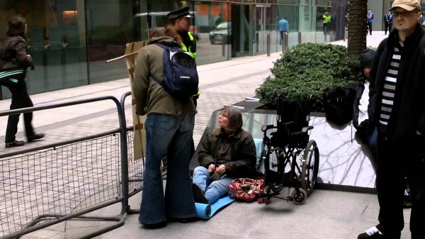 A person sits on the floor in a city centre by their wheelchair. They are being addressed by a police officer.
