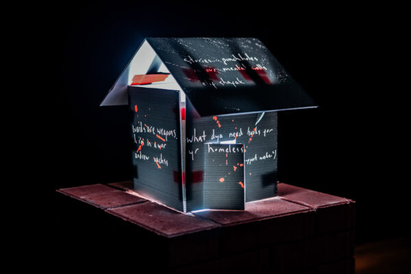 A stylised model of a house constructed from estate agent signs. The house is surrounded by darkness but is brightly lit from inside. There is handwritten wrting on the house that raads "words are weapons. & i’m in a war. the war hasn’t stopped.", "stories punch holes in our mental walls" and "'what dya need books for yr homeless" 