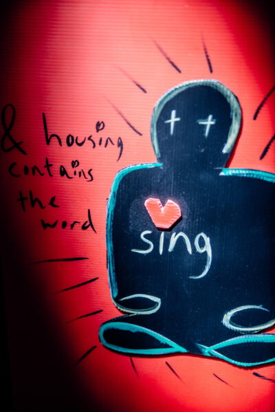The image is a mixed media collage. On the right-hand side is a figure, in black, with white crosses for eyes. The background with red. Handwritten text reads "housing contains the word sing". The first 4 words are written in black on the red background, the word 'sing' is written on the chest of the figure in white. The dot above the letter I is a read heart, and is on the centre-right of the black figure's chest.