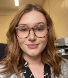 Abby, is sat in front of a plain background. She is smiling into the camera. Her hair is loose and wavy. She is wearing a grey cardigan and patterned glasses.