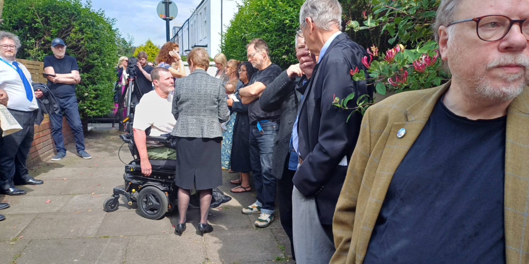 A group of about 15 people a talking and waiting. There are TV cameras and journalists. Chris Hartworth (the author) is central to the photograph in a wheelchair.