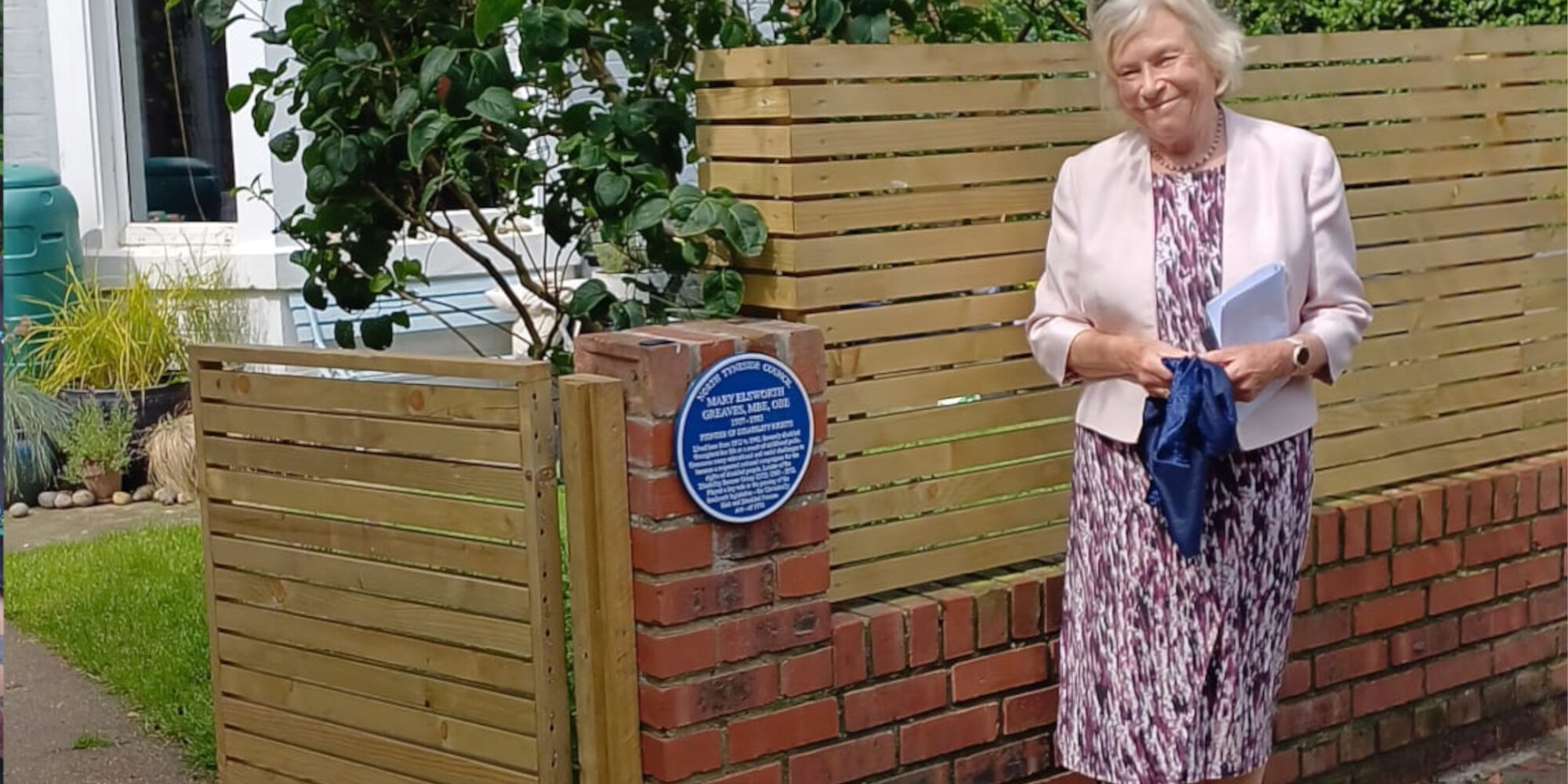 A photograph of Dame Joyce Quinn standing beside Mary Greaves' blue Plaque.