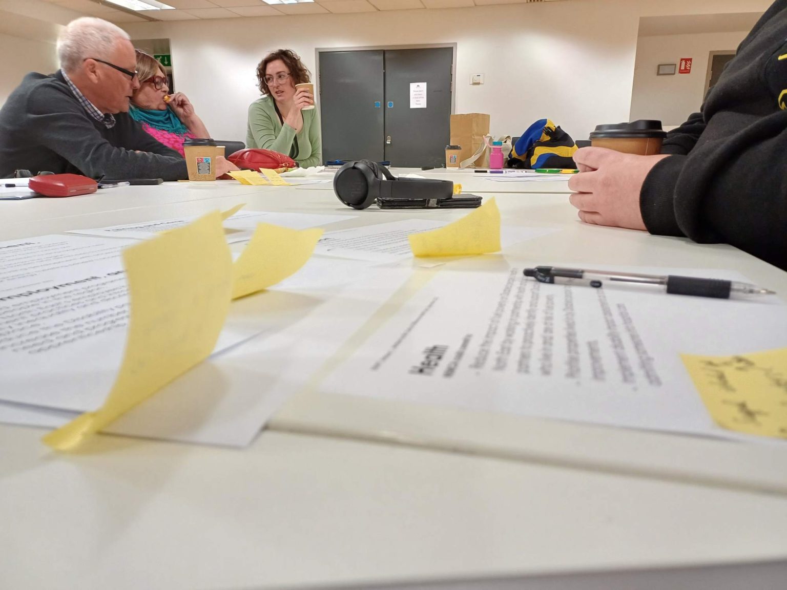 A group of people sit around a table. The table has papers and post its on that cover themes and plans for the disability devolution document.