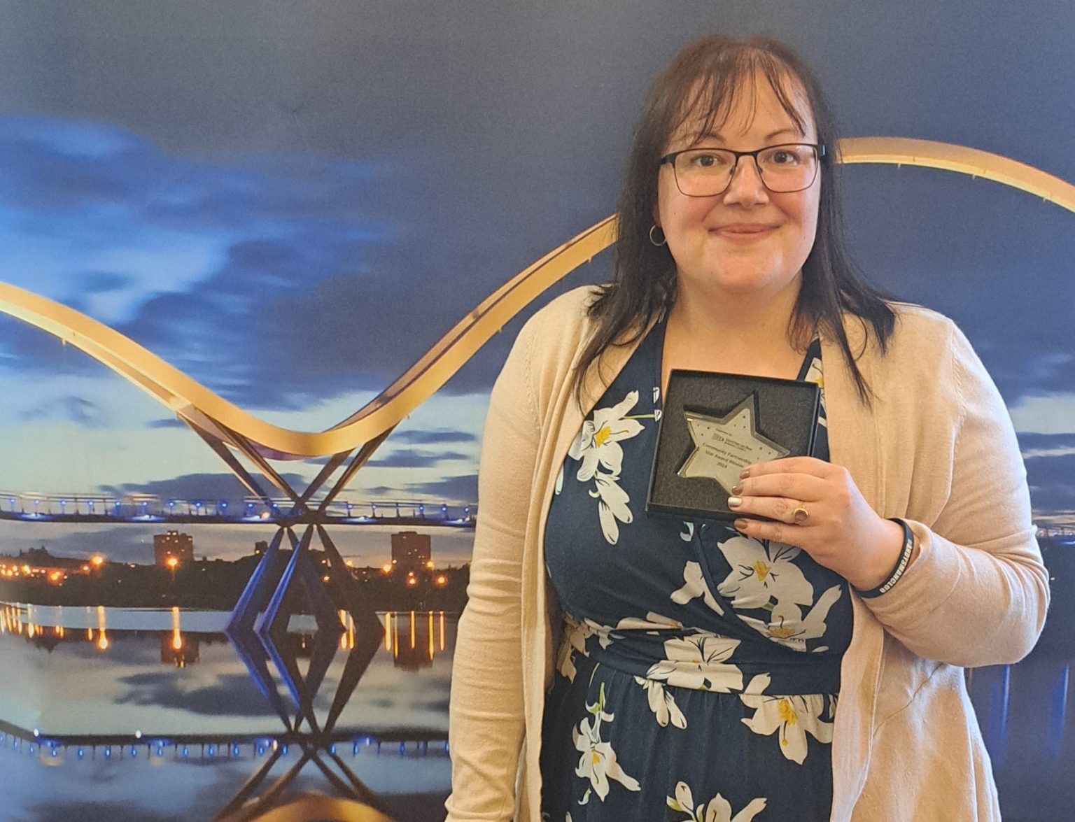 Claire has shoulder length brown hair and thick rimmed glasses. She is wearinga blue dress with large white flowers. She is smiling at the camera. She is holding an award shaped like a star and standing in front of a backdrop featuring the stockton on tees infinity bridge.