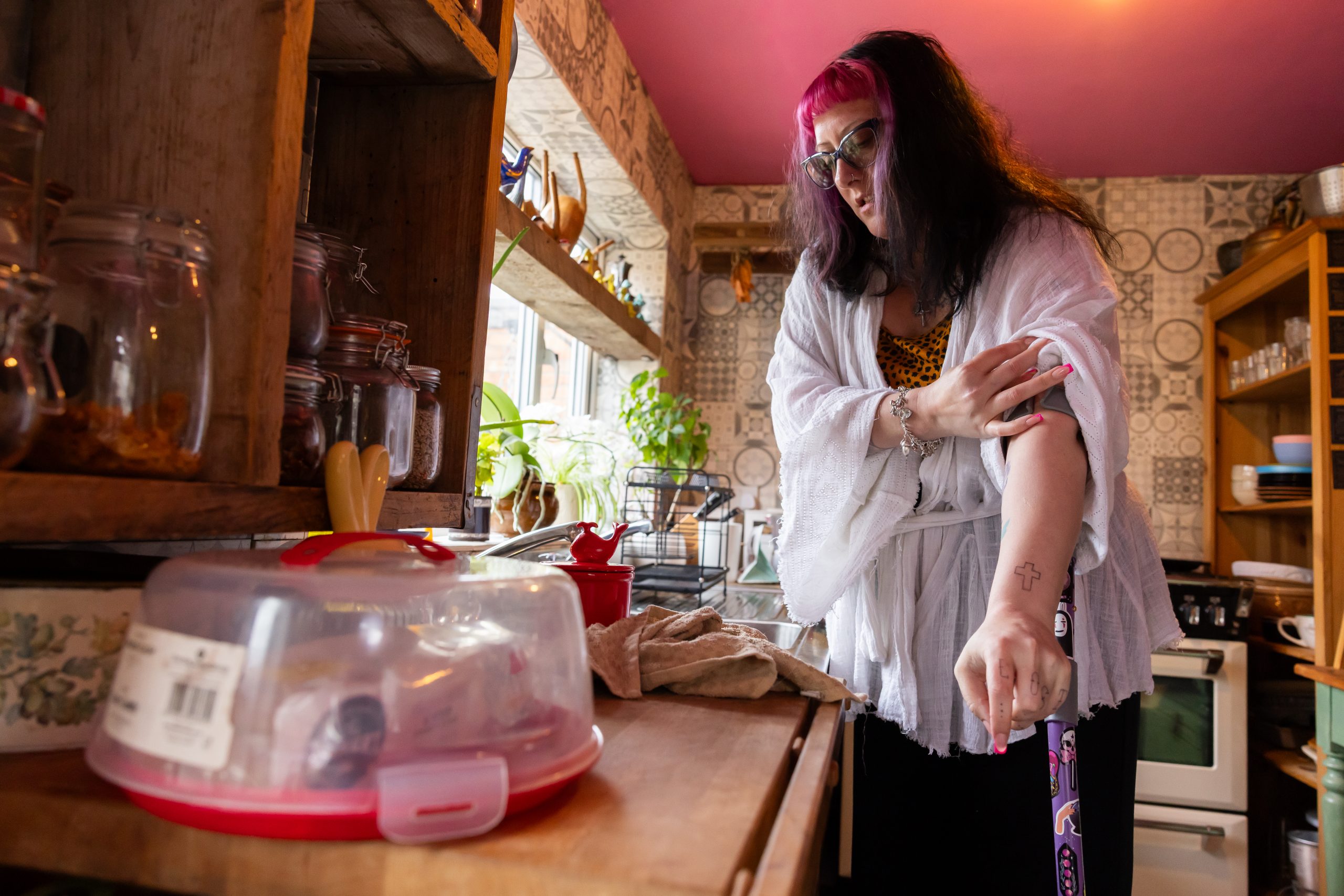 Payment for involvement. A disabled woman stands in a cosy kitchen with colourful tiles and wooden shelves. She wears glasses, a white robe-style top, and has pink streaks in her hair. She is using a mobility aid with stickers and is rolling up her sleeve to show her arm, which features a cross tattoo. Kitchen utensils, jars, and plants are visible around her.