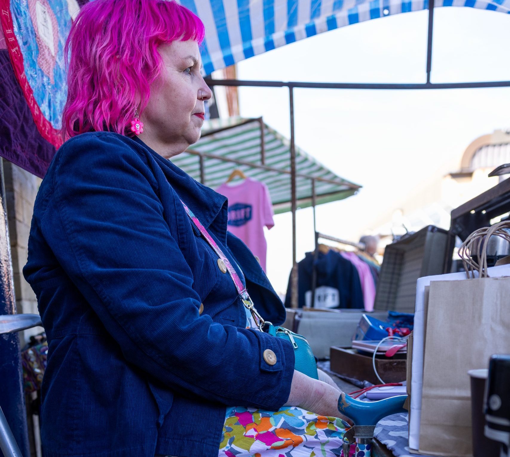 A disabled person with bright pink hair is sitting on a folding chair at a market stall. They are wearing a blue corduroy jacket, a multicolored patterned dress, and pink high-top sneakers. They are resting their hands on a colorful patterned walking stick in the foreground. The market stall includes various items for sale, with a blue-and-white striped canopy overhead. The person is looking to their left with a calm expression.