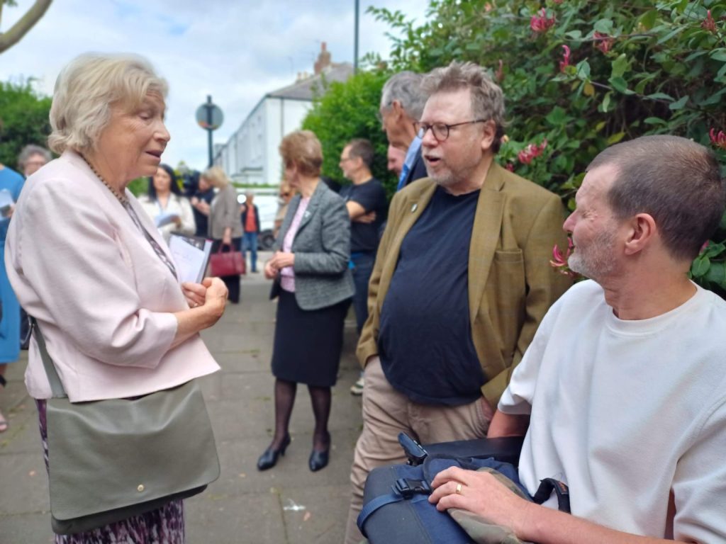 A group of people are gathered outdoors on a sidewalk, engaging in conversation. In the foreground, an older woman wearing a light pink jacket and carrying a green bag is speaking to two men. Colin Cameron is wearing glasses and a bearded wearing a brown blazer and black shirt, he is beside another man, who is seated in a wheelchair, wears a white shirt. In the background, more people are standing and talking near greenery and residential buildings.