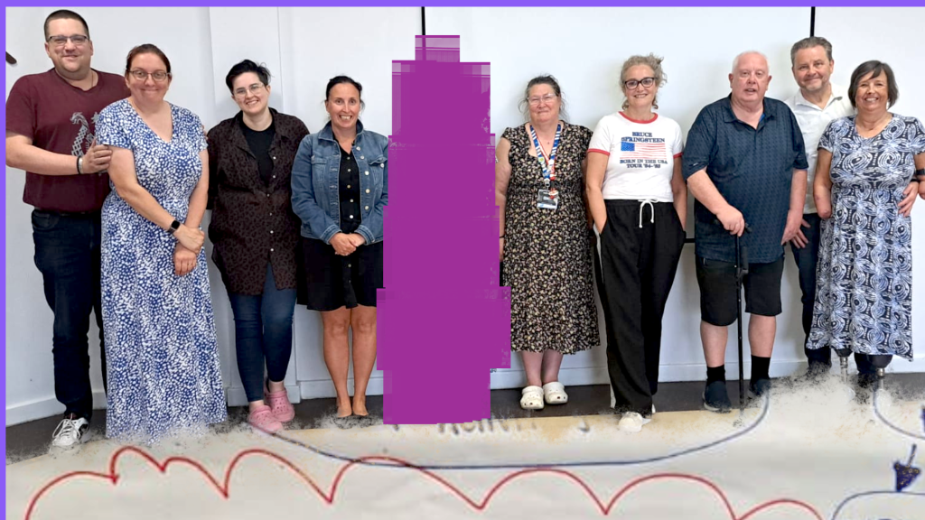 Hartlepool Disability Rights: A group of twelve people stand together, smiling, in front of a white wall at an indoor event. They are of varying ages and genders, some using mobility aids.