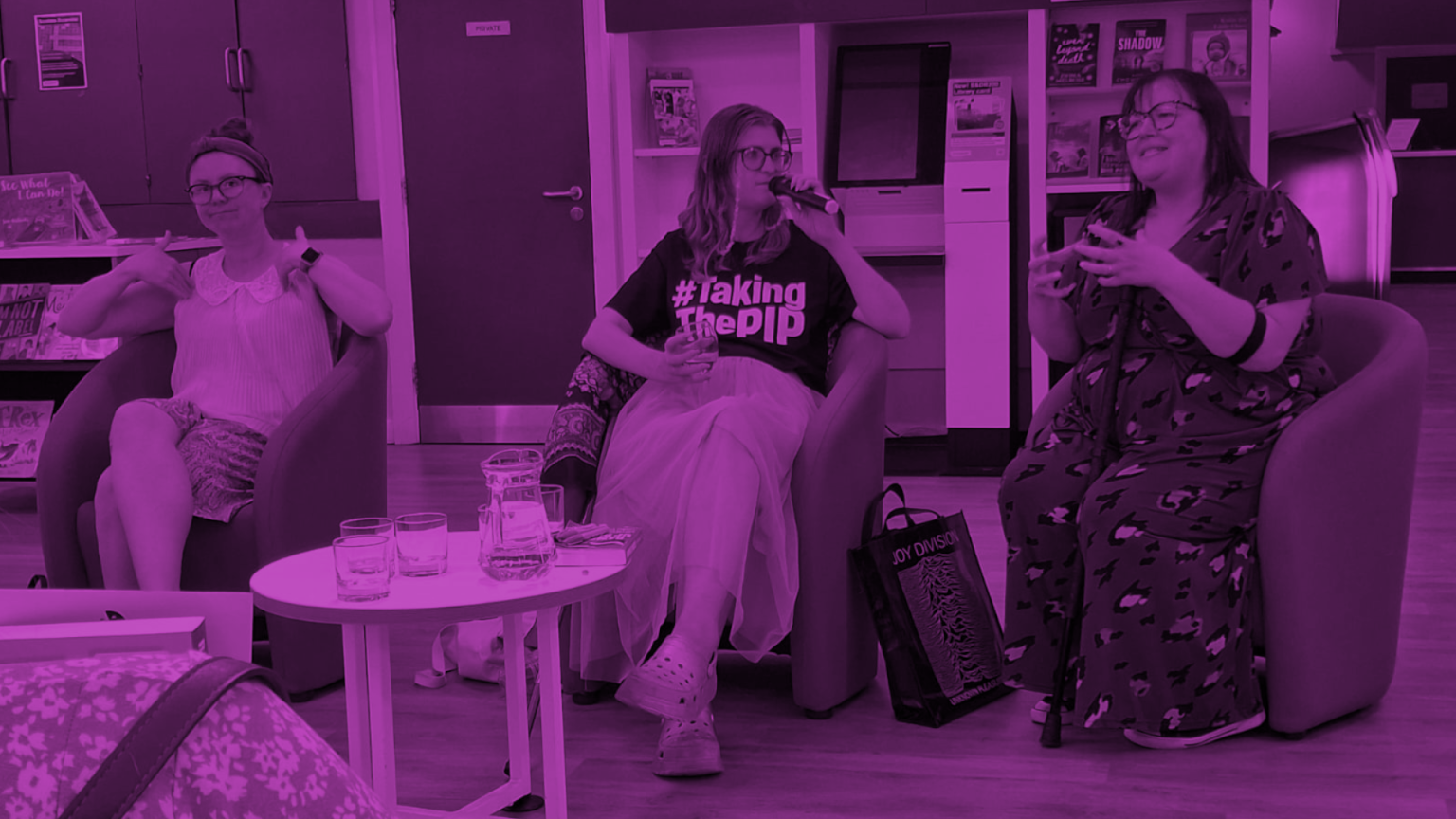 Ramping Up Rights: Teesside. Three disabled speakers sit in armchairs during the “Ramping Up Rights” event at Stockton Library. The person in the centre holds a microphone and wears a T-shirt that reads “#TakingThePIP.” A Joy Division bag is on the floor nearby. The setting is relaxed and informal, with bookshelves in the background and water glasses on a table in front. The image is tinted purple.