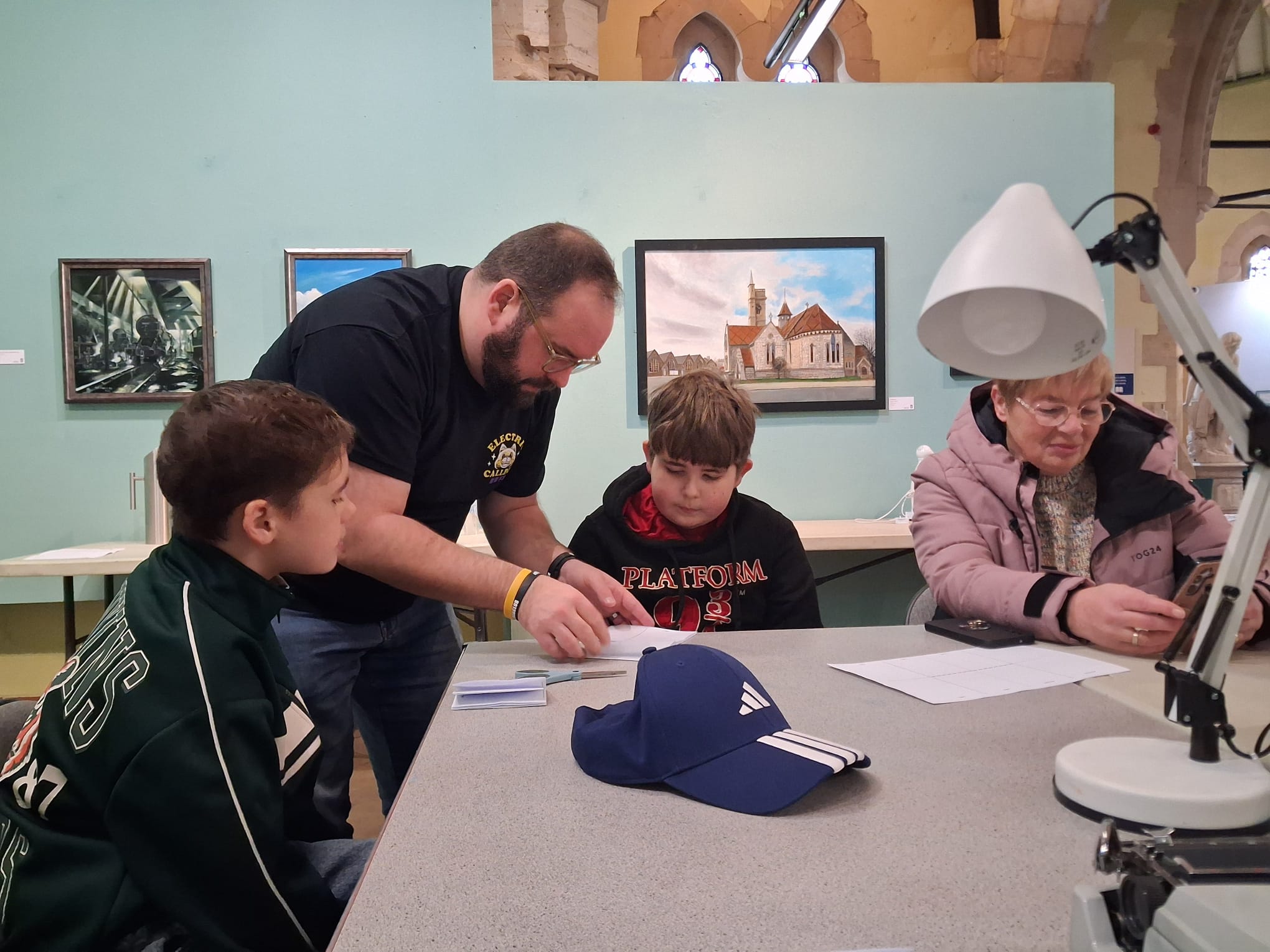 Zine making in hartlepool, Two young people are seated looking with concentration at their work infront of them, an adult stands next to them leading over slightly. the background is of artwork and photography in an art gallery setting