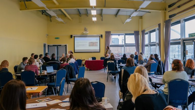 Cleveland Police Hate Crime Strategy Launch: Attendees seated at round tables in a bright community hall, facing a presenter standing beside a projected presentation screen at the Cleveland Police Hate Crime Strategy launch