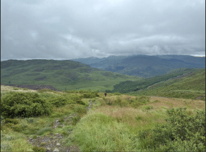 History Workshop Disabled People North East: A wide view of rolling green hills and mountainous landscape beneath a cloudy sky, with a narrow rocky footpath winding through tall grass in the foreground and a lone walker in the distance.